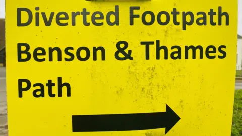 Bright yellow sign showing footpath diversion of Thames Path to Benson with a large, black arrow.
