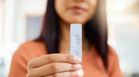 Getty Images A woman with a positive Covid test after checking it at home. She holds a white rectangle of plastic with indicators to show if the test is positive. She has long dark hair past her shoulders and wears an orange top.