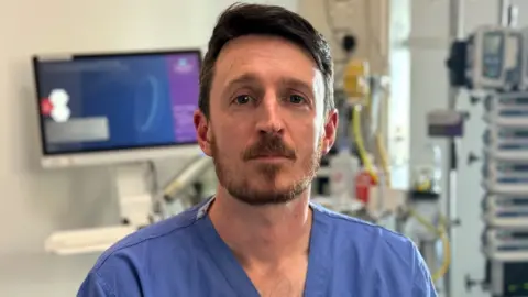 A man sitting in a medical theatre looks into the camera. He has short brown hair and a brown beard. He is wearing blue medical scrubs.