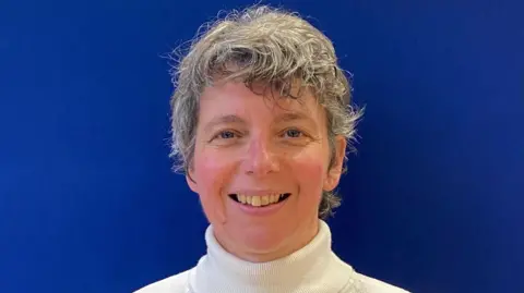 Hope Works Kirstie Cook standing in front of a blue wall. She is wearing a white turtle necked jumper and is looking straight at the camera and smiling. She has short fair hair. 