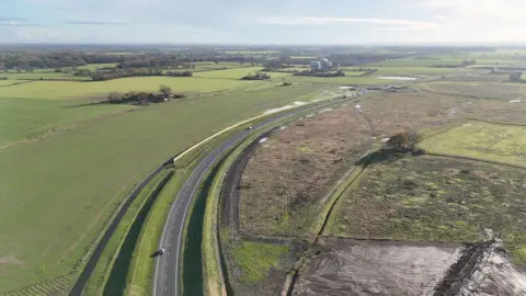 BBC/Qays Najm A drone picture of the Long Stratton bypass which has grey tarmac and bends through green fields. There are only a handful of vehicles on the road. The sky is blue and the sun is shining
