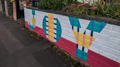 A colourful piece of wall art outside a residential property. It says JOY and has been decorated with colours of red, green, blue, yellow and pink. 