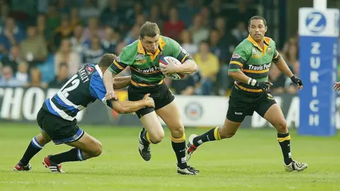 Getty Images A man in a green and yellow rugby shirt carrying a rugby ball runs past a man trying to tackle him. 