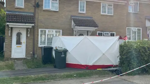 Helen Mulroy/BBC A white tent outside the entrance to the property, which also has two black wheelie bins out the front. Red and white tapes off the entrance to the house.