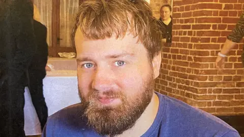 Tom James is looking directly at the camera in a close-up photograph. He is indoors. Behind him is a bare brick wall and several people, and two tables with white tablecloths on.
