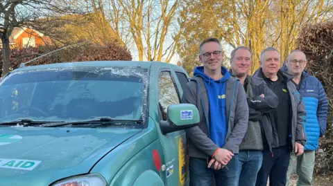 Four men, Alistair Gray, Paul Earnshaw, Fergus Ramsay and Craig Wrightson, are standing next to a green 4x4 humanitarian air car which has frost on the windows. The men are smiling at the camera and are wearing coats. 