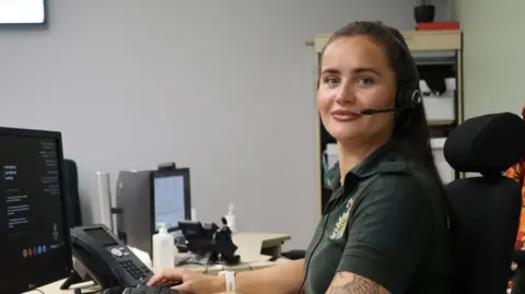Nicole Douglas is sat at a desk looking at the camera. She has long, dark hair and is wearing a headset and a green shirt. There is a telephone on the desk as well as various monitors.