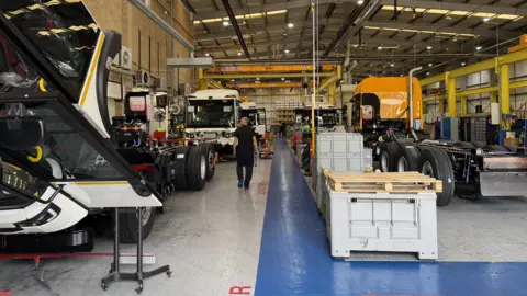 The inside of a warehouse with part of lorries in them, including wheels and cabs. There are wooden pallets around and a blue walkway on the floor