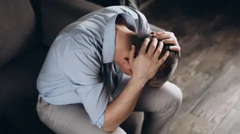 A man in despair with his head in his hands. He is wearing a blue shirt and light trousers. We cannot see his face but he has short, cropped dark hair.