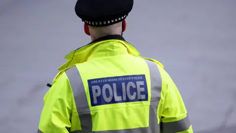 A male police officer from Greater Manchester Police on duty in the city centre. They are facing away, wearing a yellow high-vis jacket and a black police hat.