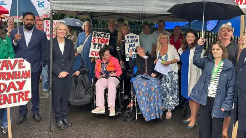 Hazel Howe A line of people standing under a gazebo. Two of them are in wheelchairs. Some are holding umbrellas. Some are carrying placards which say "Bucks stop the cuts" and "Don't close my day centre".