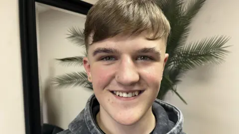 A teenage boy with brown hair is smiling at the camera,
He has a number of facial piercings