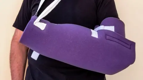 Getty Images A man in a black T shirt stands with his arm in a purple foam sling