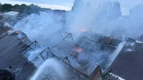 A large warehouse roof on fire as it is hosed down from above