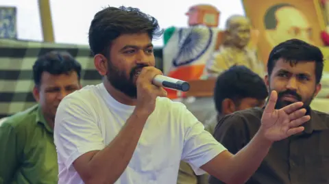 EPA Abhijeet Dipke wears a white shirt, holding a microphone and speaking