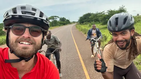 Contributed A cyclist takes a selfie while riding along a deserted road. He has a silver helmet on, glasses and a red top. Next to him on the right is another cyclist with his thumb up, wearing a grey helmet, light brown cycling jersey and black cycling shorts. Behind them both are two more cyclists wearing normal pedestrian clothes but without helmets. 
