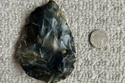 Emma Witten The black and brown flint hand axe which has chisel marks on it next to a 20p piece for scale