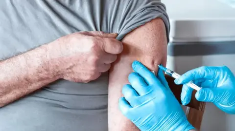 A man receives as injection in his arm from someone wearing blue surgical gloves.