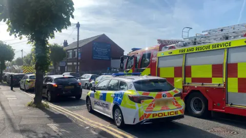 A police car is parked on the side of a residential road. A red fire engine is parked in the middle of the road with cars parked on either side of the street. Further up the road we can see an ambulance and several more fire engines.
