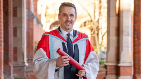 Gary Neville is smiling as he stands in between a series of tall red brick columns. He is holding an honorary degree and is wearing university graduation robes.