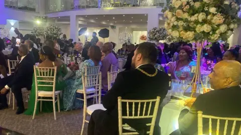 Nana Ama Ofori-Atta The audience at an awards ceremony sitting at round tables looking towards the front. They are mostly in formal dress and there are tall floral displays on the tables.