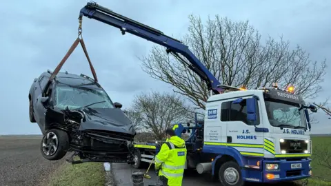A van with a crane holding up a smashed up car. The van has neon stripes and "J.S. Holmes" and "recovery" on its front and side. The damaged car is black has a crunched front bonnet and heavily cracked window screen. They are on a road with fields and trees on either side. A man in a high-vis coat is facing towards the smashed car. 