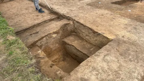 Tony Fisher/BBC A neatly excavated pit reveals distinct soil layers and stepped sections cut into the ground. The edges are sharp and precise, showing careful archaeological methods. An archaeologist stands nearby using a tool to clean and define the trench. The surrounding cleared surface and marked areas indicate an organised and ongoing excavation.
