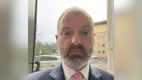Matthew Pitts A man, pictured from the shoulders up, with short grey hair and a grey beard. He is wearing a white shirt, pink tie and navy suit jacket. He is standing in front of a window. 
