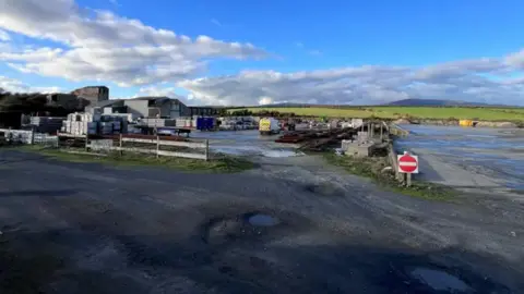 Grey gravel with fences, boxes, puddles and a red 'no enter' sign are in the foreground with a green field, hills and a blue and cloudy sky in the background