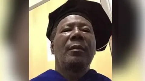 Submitted Dr Morris is pictured in a blue University graduate gown and is wearing a black graduate hat. 