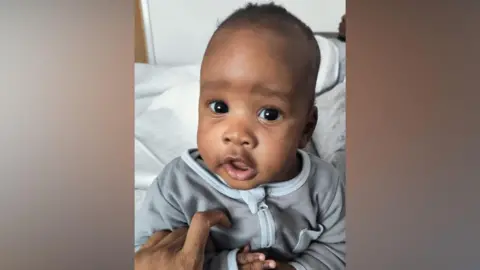 A young baby boy with short dark hair looks towards the camera. He is wearing a grey zip-up babygrow and is being held by an adult whose hand is visible.