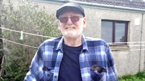 George Mackay standing in the back garden of a grey house. He is wearing a cap, a blue checked shirt, dark blue t-shirt and sunglasses, and has a white beard.