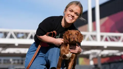 Ellie Simmonds, with light hair, pulled back behind her head, leans slightly to one side while holding her arms around a dog as she kneels. She wears a black T-shirt and blue denim jeans. The dog she holds has light brown fur with a strip of white on its chest.