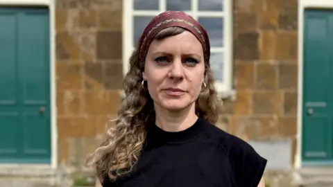 Wood stands with long curly hair, a head scarf and the top of a black peak in front of a small church of orange brick.