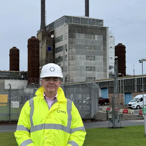 Richard Murray A man in a high visibility jacket and white hard hat stands in front of a former nuclear plant