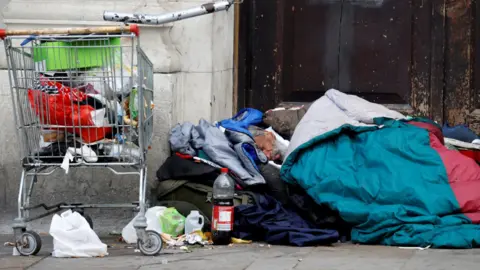 Homeless person sleeps in a door in a sleeping bag near a shopping trolley.