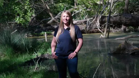Shaun Whitmore/BBC Ursula Juta is standing by the flooded woodland, beside one of the beavers' dams. She has long blonde hair and is wearing jeans and a navy t-shirt.