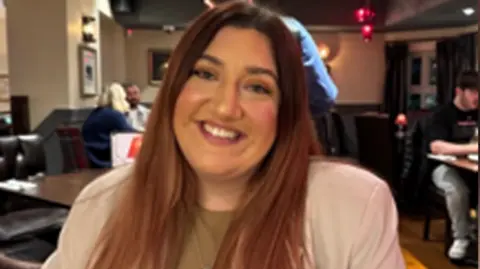 A woman looking at the camera. She has long brown hair and smiles at the camera. She is wearing a beige blazer with a brown top underneath.
