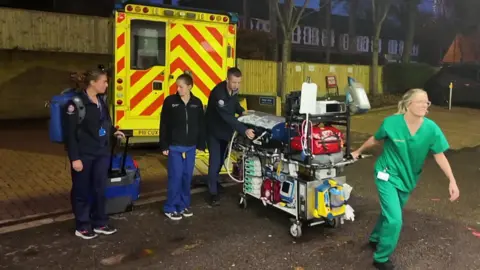 Team of medics leaving an ambulance. There are four figures. Two women and a man are wearing navy blue jackets and trousers. There s a woman wearing green scrubs pulling a trolley full of equipment. She has long blonde hair in a pony tail and is wearing glasses. The ambulance is parked next to a wooden fence and trees. 