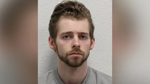 Custody picture of Joshua Michals, wearing a grey sweat top and he has light short brown hair combed back and a beard.