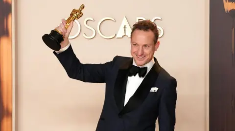 Lol Crawley poses in the press room during the 97th Annual Oscars at Dolby Theatre in Hollywood, California. He is wearing a black tuxedo with a black bow tie and is holding his Oscar up in celebration.