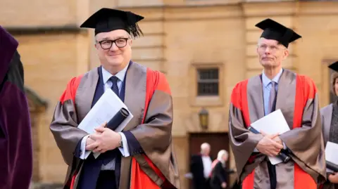 PA Media A man with glasses and wearing a blue suit, white shirt, blue tie, grey and red silk robe and a graduation cap carries documents and a black tube as he leaves a theatre following an honorary degree ceremony at the University of Oxford. Behind him is a man wearing glasses, a dark suit, pale blue shirt and tie, a grey and red silk robe and a graduation cap. He is also carrying documents and a black tube. There are other people in robes blurred in the background. 