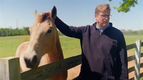John Miller pets his golden-coloured horse while standing by a wooden fence in his field