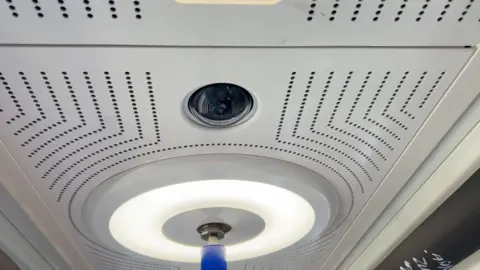 A camera is in the roof of the train and there is a circular light around a blue pole