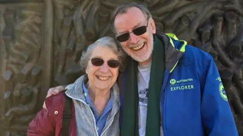 Jenny is on the left, standing with her husband John on the right with his arm around her. Both are smiling. She has short grey hair and he has thinning grey hair and a short beard. Jenny is wearing a red jacket and John a blue jacket and green scarf.