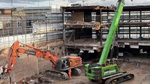 West Northamptonshire Council A building site, showing red and green machinery, with buildings being demolished around it. 