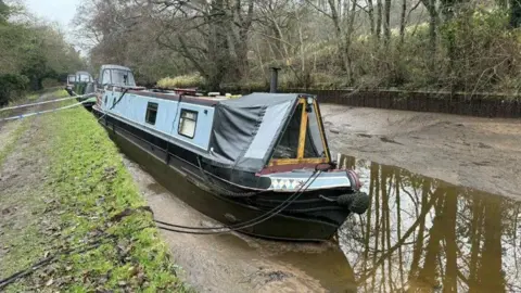 Phil Johnson A canal boat tilted slightly to the right, sitting on the bottom of a canal that has been drained completely, apart from a small amount of water.