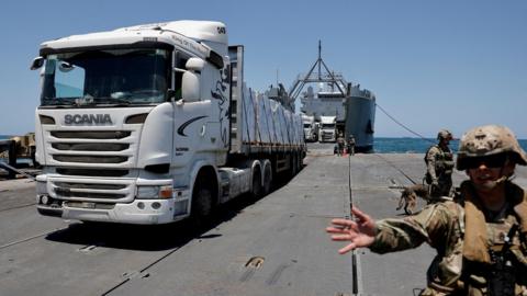 A US serviceman gestures as a lorry transports aid delivered via the US military's floating pier off the coast of Gaza (25 June 2024)