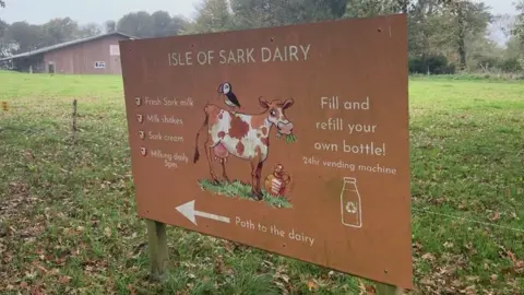 A rustic large sign stands in a grassy field near a wooden with what looks to be a slate roof, surrounded by leafy trees. The sign reads “Isle of Sark Dairy” in bold letters and features playful illustrations of a black-and-white cow with a puffin perched on its back, a glass milk bottle, and a traditional metal milk churn. Below the title, the sign lists offerings: fresh Sark milk, milkshakes, Sark cream, and notes that milking occurs daily at 5pm. It encourages visitors to fill or refill their own bottles using a 24-hour vending machine. An arrow points left with the words “Path to the dairy.”