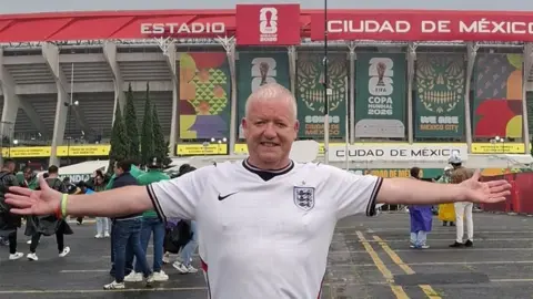 Paul Clegg in a white England shirt standing outside the Azteca Stadium in Mexico with his arms stretched out to the sides. He has short white hair 